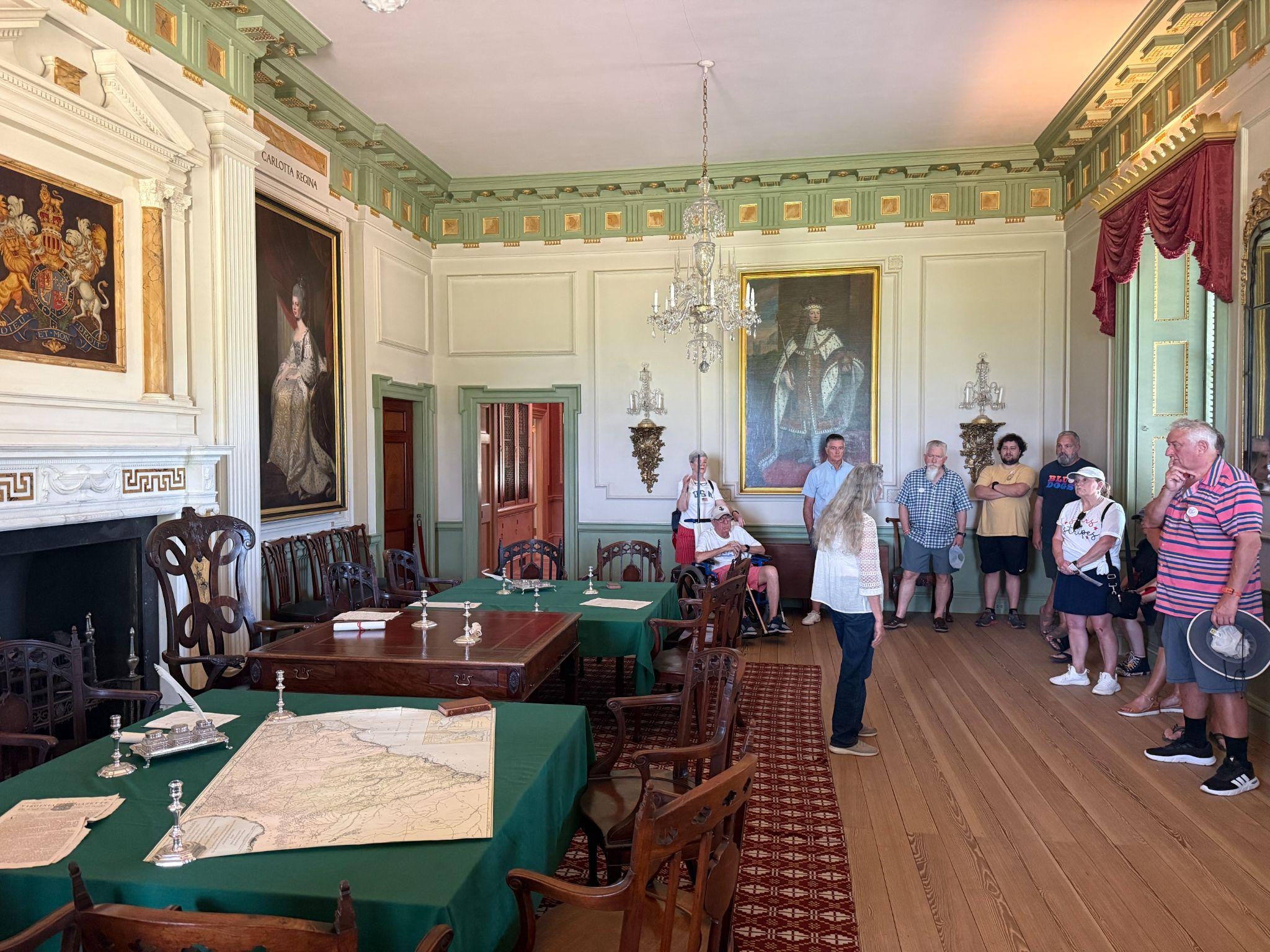 Interior council chamber at Tryon Palace in New Bern with royal portraits, a chandelier, and a guided tour group